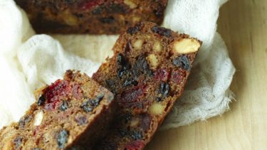 Fruitcake loaf with two cut slices sits on white sheet