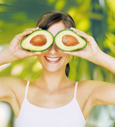 Young woman holding halved avocado over eyes, smiling, close-up
