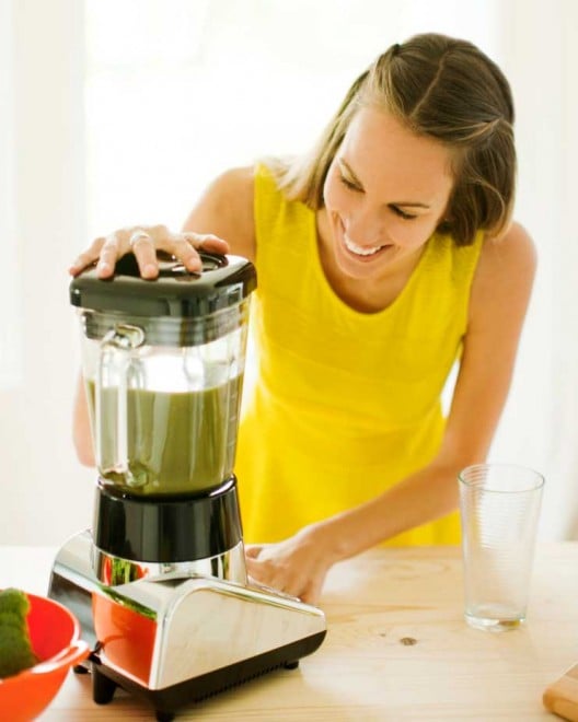 Woman wearing a yellow shirt uses a blender full of vegetables. Woman is looking down and smiling at the blender. An orange bowl of broccoli is visible in the foreground. And empty glass sits on the counter next to the blender.