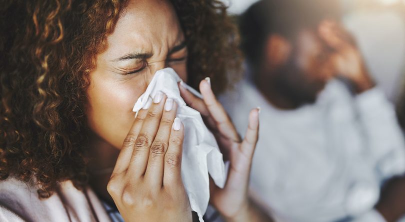 Flu explained: Shot of a young woman blowing her nose with her boyfriend in the background