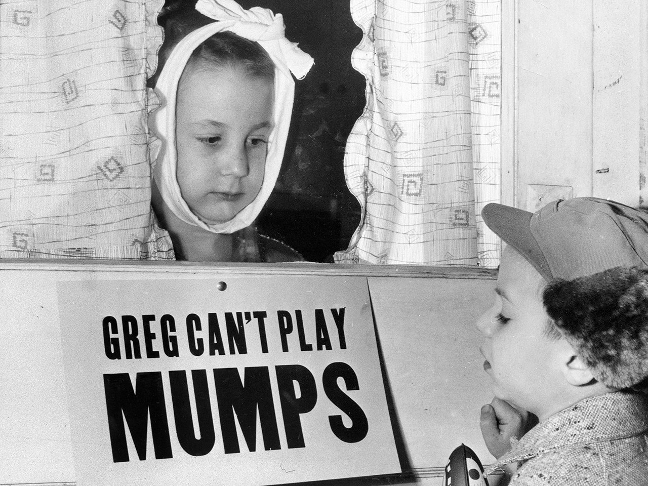 In this Jan. 16, 1957 file photo, Jon Douglas, 6, right, visits his friend, Greg Cox, standing behind a sign warning he has mumps, on the door of his home in Altamont, Ill. Fifty years ago, mumps was once a childhood rite of passage of puffy cheeks and swollen jaws. That all changed with the arrival of a vaccine in the late 1960s, and mumps nearly disappeared. But in 2017, the U.S. is in the midst of one of the largest surges in decades. Photo, AP.