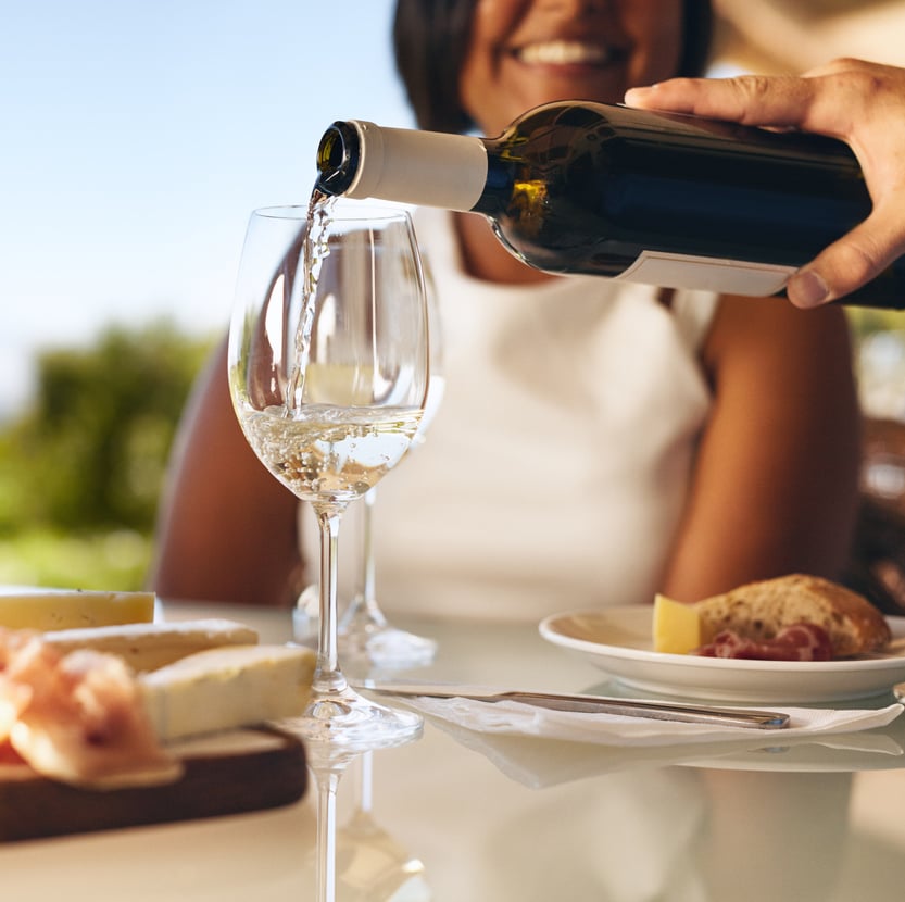 Hands of a man pouring white wine in two glasses from bottle with a woman smiling in background at winery. Focus on glasses and wine bottle.