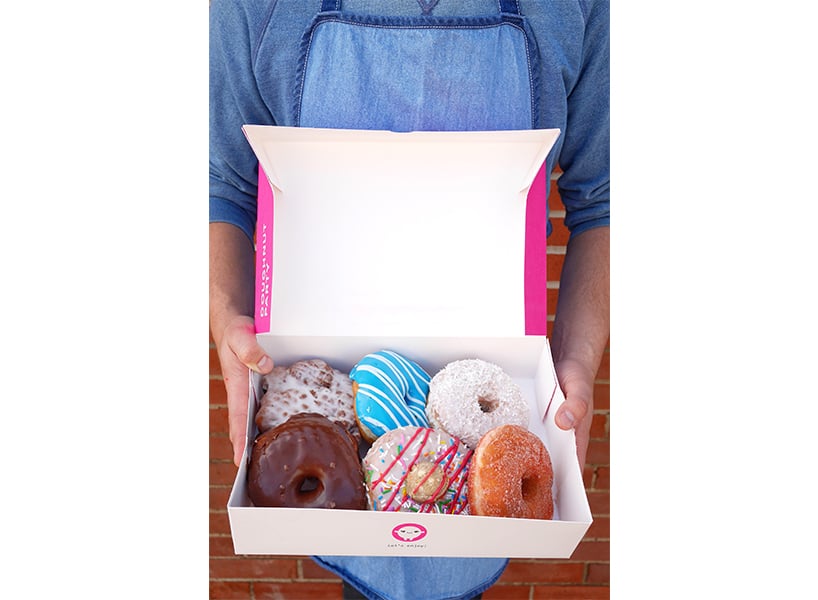 A man in a blue apron holds an open paper box filled with six brightly coloured doughnuts from Donut Party in Edmonton.