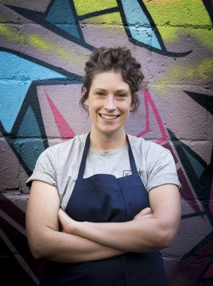 Chef Alexandra Feswick in navy apron, standing with arms crossed in front of a graffiti wall