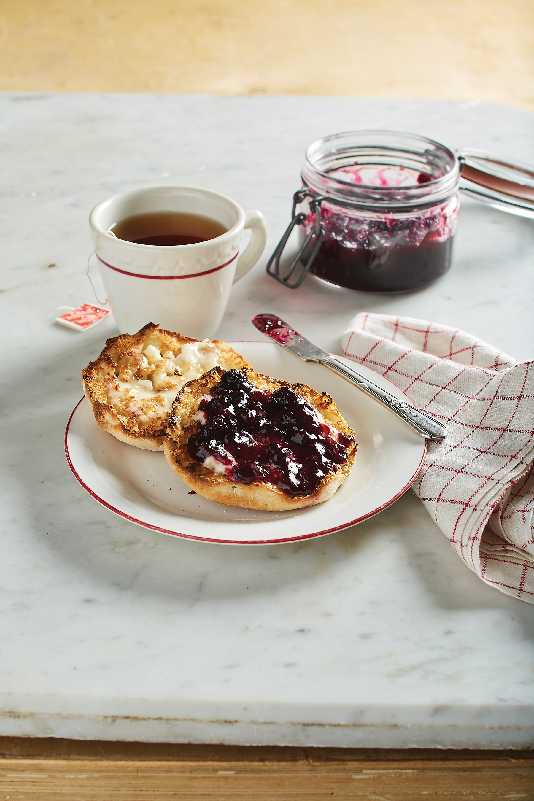 Scone covered in blueberry jam on plate in fromy of mug of tea and jar of jam
