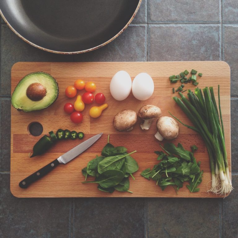 A cutting board with avocado peppers, tomatoes, eggs, green onions, spinach and herbs prepped on it