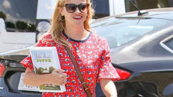 Reese Witherspoon walking down the street in jeans and a red shirt carrying a novel