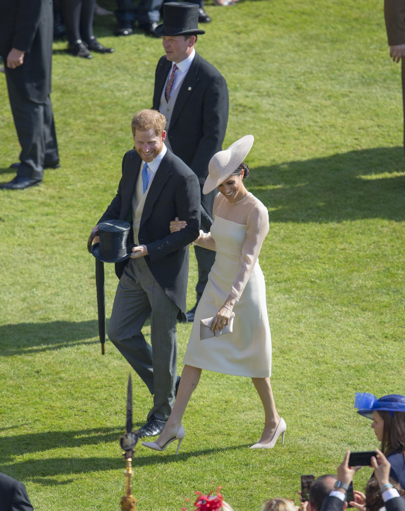 Prince Harry and Meghan Markle at a garden party. Markle is wearing a hat and pantyhose