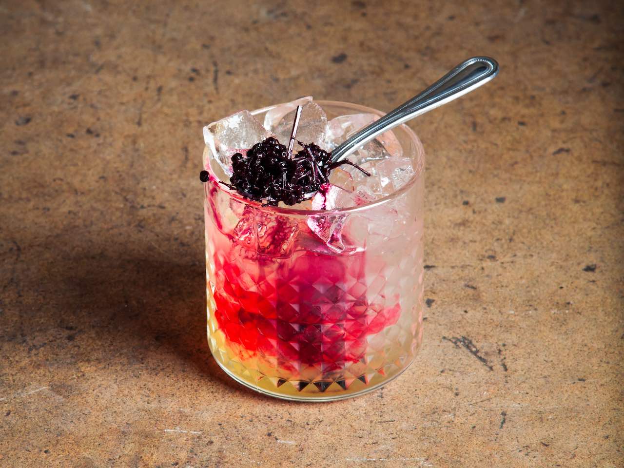 Pink cocktail in glass topped with berry and spoon.