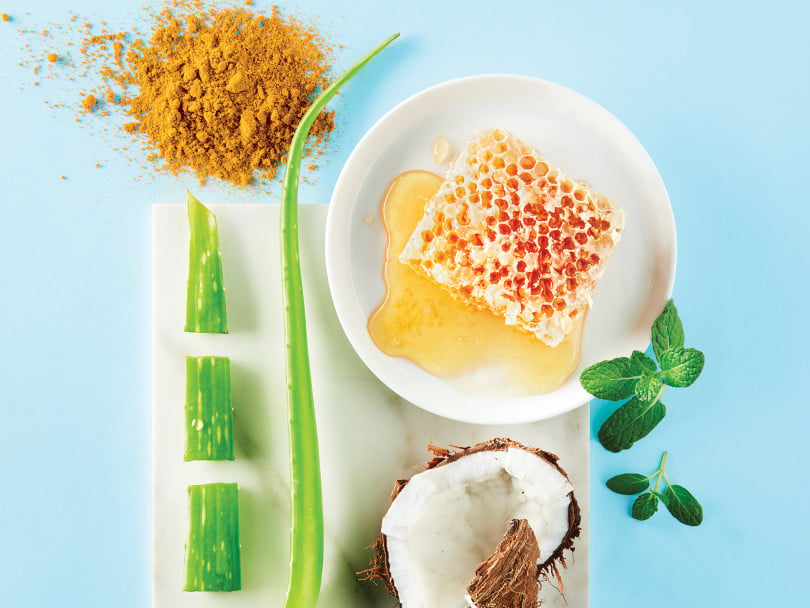How Clean Is Your Beauty? Tumeric, aloe, honeycomb in bowl, coconut chunks, and mint leaves displayed on blue background