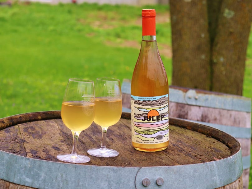One bottle and two glasses of Negondo's orange wine, Julep, on a barrel against a grassy background.