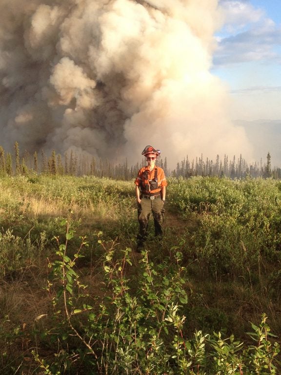 Julie Stankevicius stands in field in firefighting gear as smoke goes up behind her