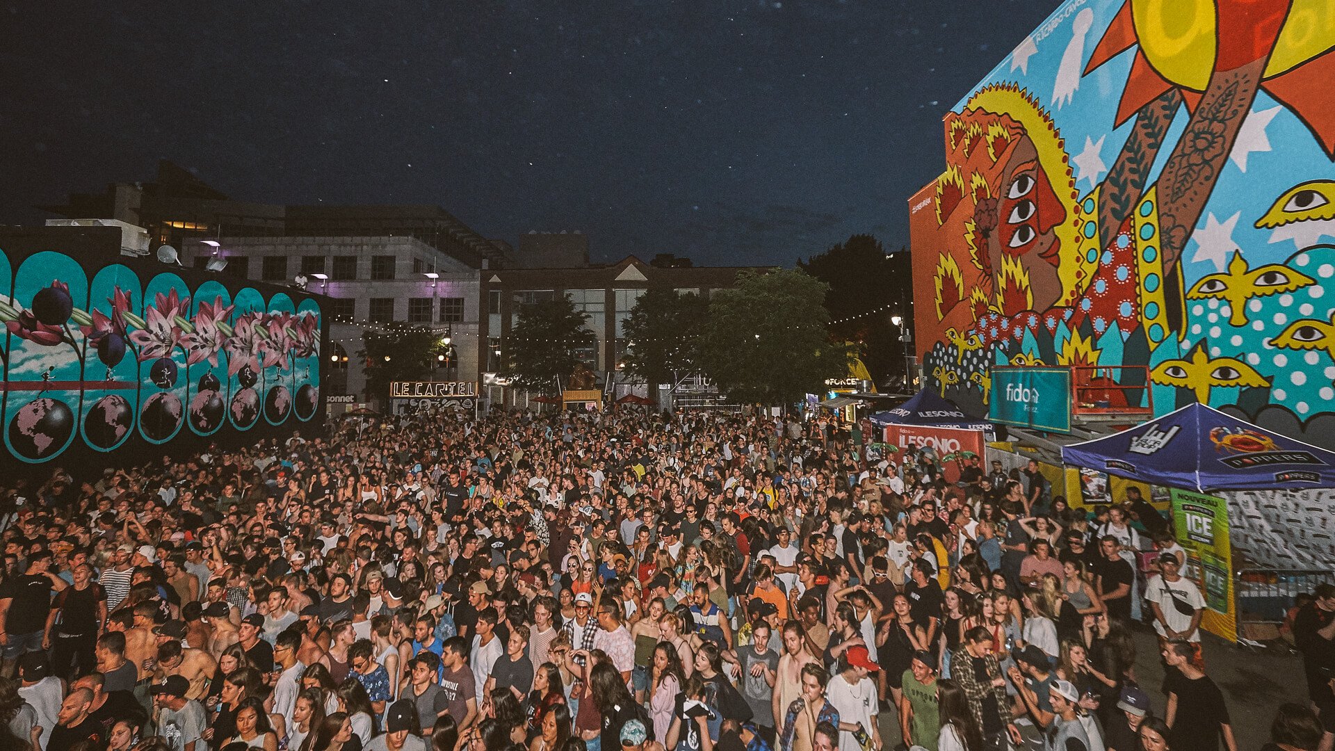 Photo of Mural Festival at night. There are two murals on either side, with a large crowd of festival-goers in the middle.