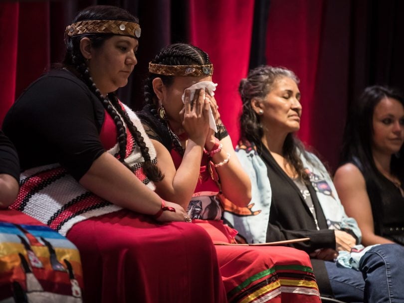 Four women sit in a row, one of them is crying into a handkerchief and two are dressed in traditional Indigenous clothing. They are responding to the report on the National Inquiry into Missing and Murdered Indigenous Women and Girls, along with other Indigenous women and allies in Vancouver, on Monday June 3, 2019