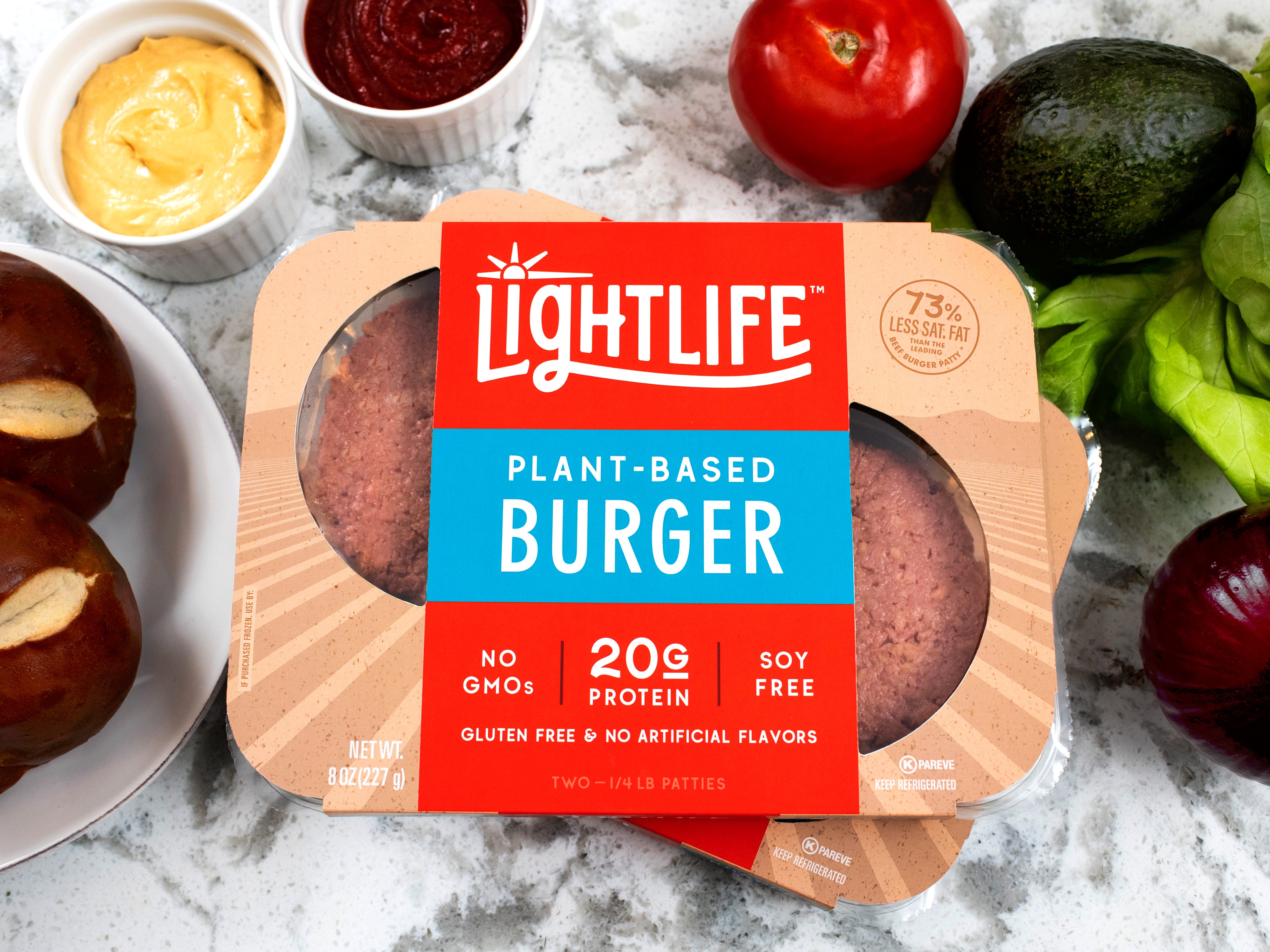 Uncooked Lightlife plant-based burgers in red, blue, and brown packaging on a marble counter surrounded by vegetables.
