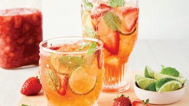 A glass pitcher and short glass sit filled with light orange bubbly liquid with floating strawberries and mint leaves, next to a mason jar filled with mushy-looking strawberries