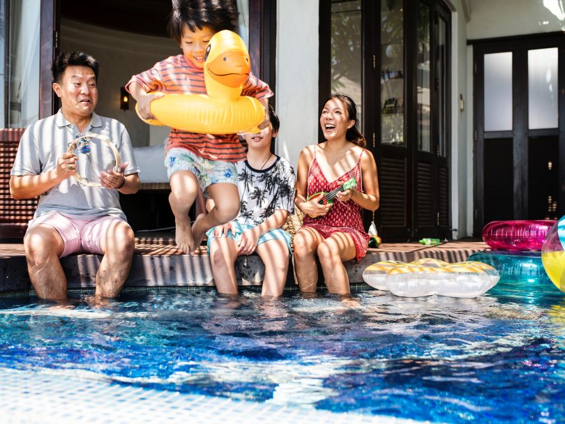 Family of four sitting by the pool. A young boy with a duck floaty toy runs into the pool as his family looks on.