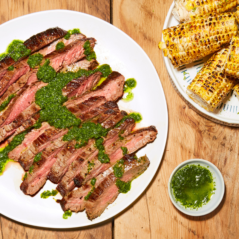 Seared flank steak on a white plate next to a plate of grilled corn.