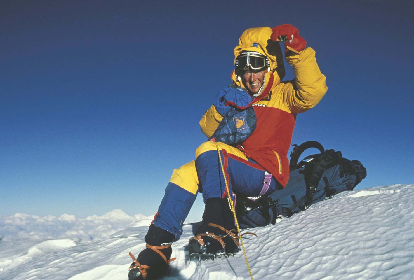A woman in snow gear and crampons sits atop a snow hill squinting at the camera