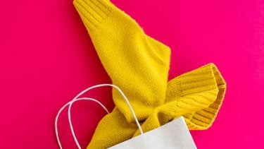 A yellow sweater peaking out of a white paper bag against a pink background.