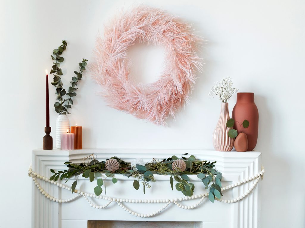 a white mantel and wall decorated with a pink wreath, maroon and burnt-orange candles and vases, greenery and wooden ball chain
