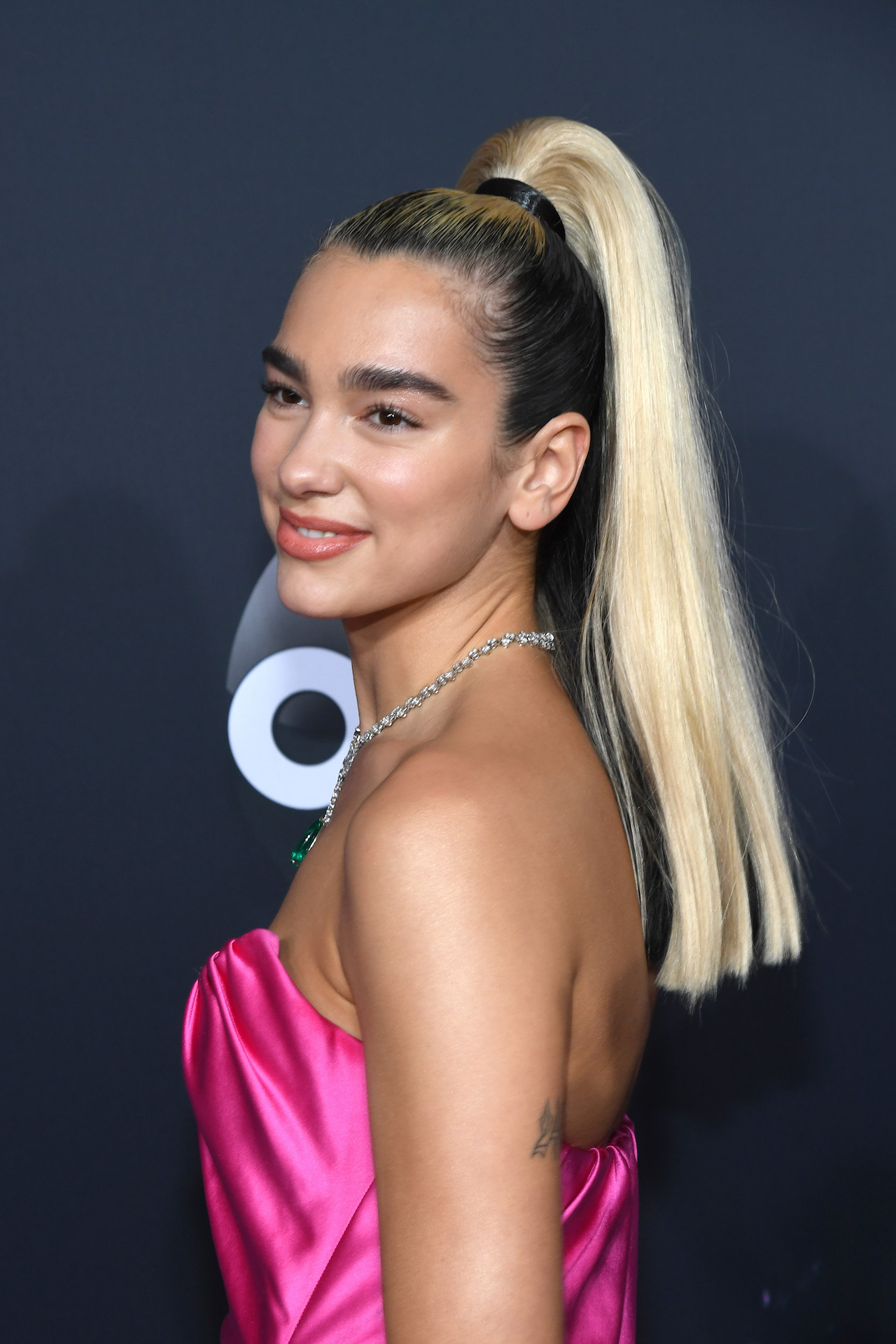Dua Lipa wearing a bright pink satin dress with her long blonde hair in a high ponytail and dark roots showing