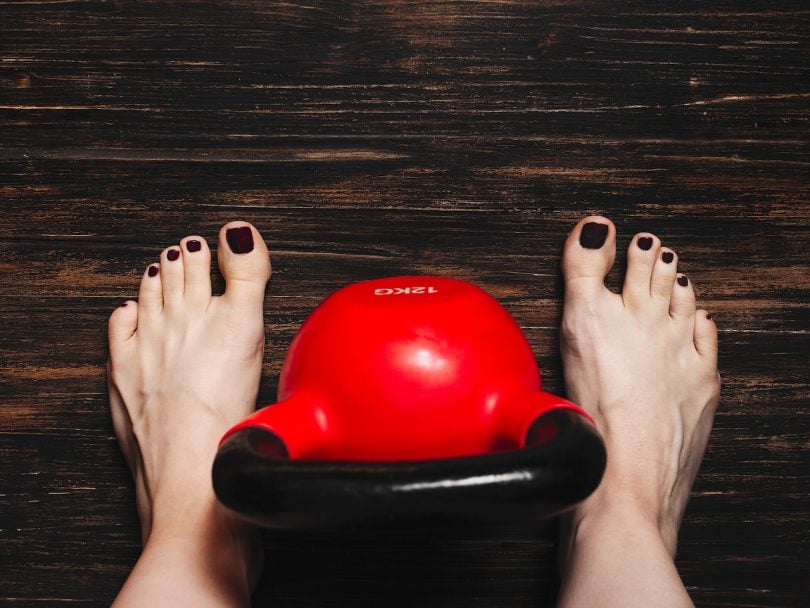 Woman standing bare foot with red kettlebell between her legs on dark wooden floor, view from above