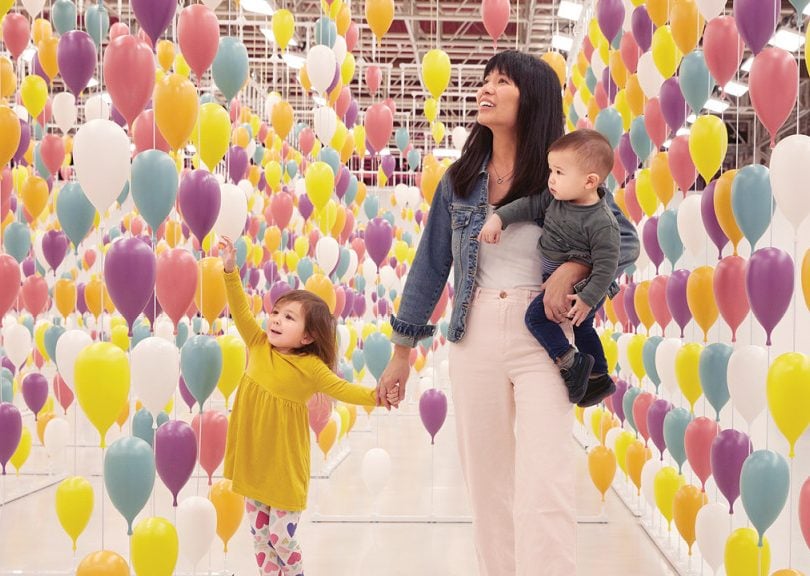 a mom and her two kids walk through a mirrored hallway covered in balloons