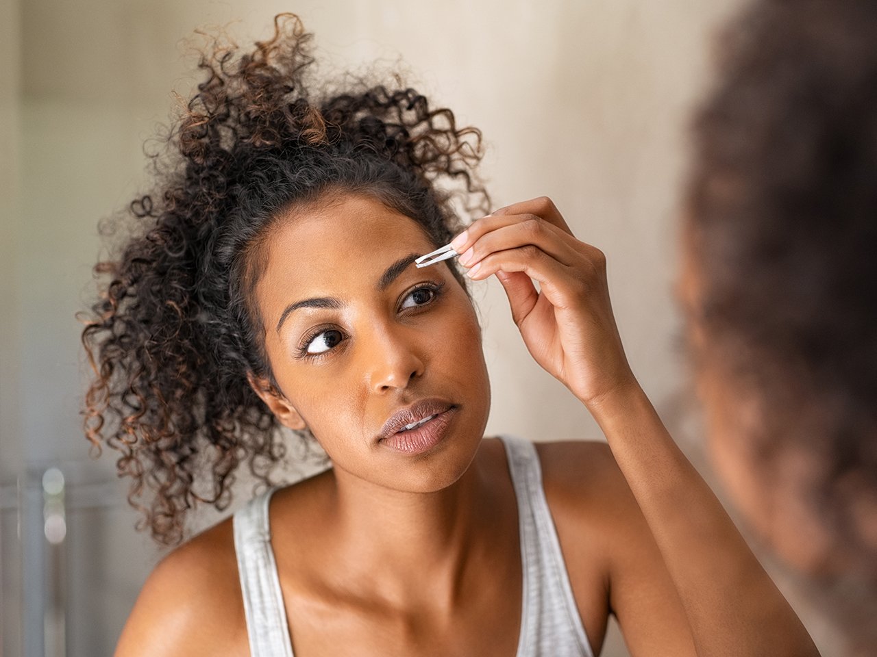 A woman plucking her eyebrows in the mirror to illustrate an article about how to do your eyebrows at home.