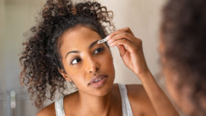 A woman plucking her eyebrows in the mirror to illustrate an article about how to do your eyebrows at home.