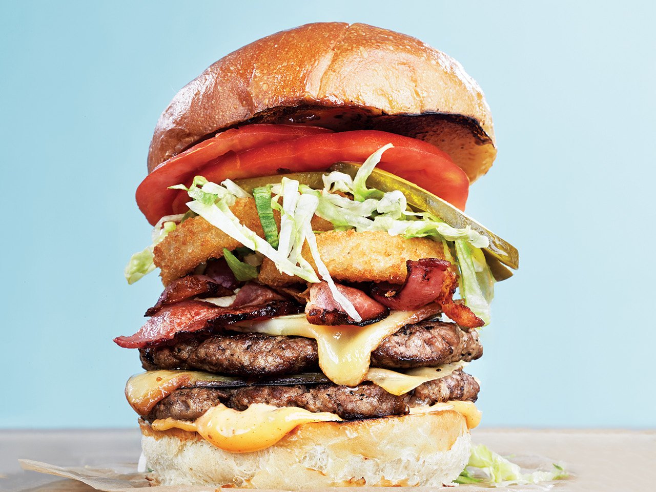 Burger stacked with beef patties, sauces, bacon, onion rings, lettuce, pickles and tomatoes.