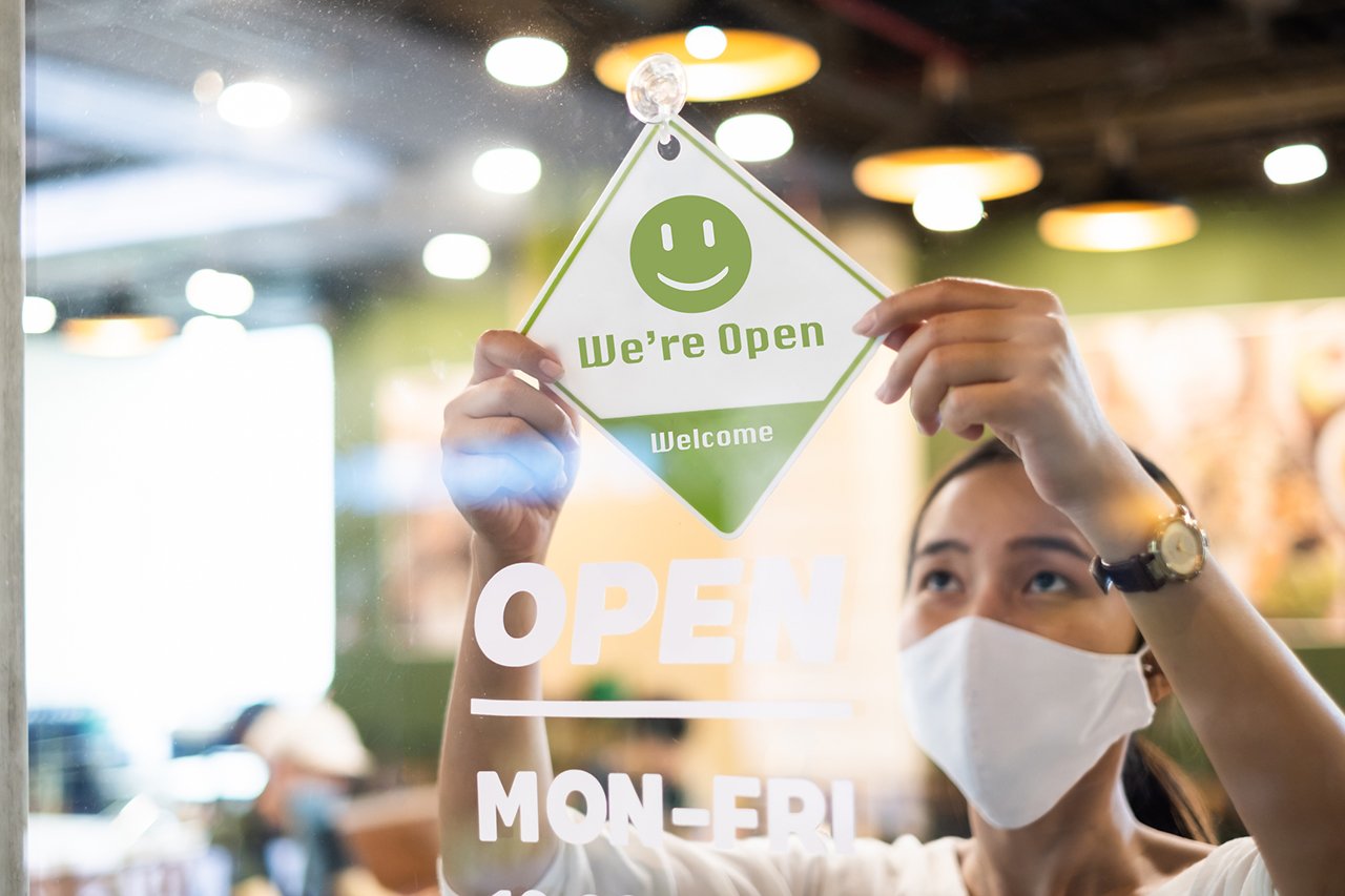 A woman wearing a face mask puts a sign saying "We're Open" in a store window 