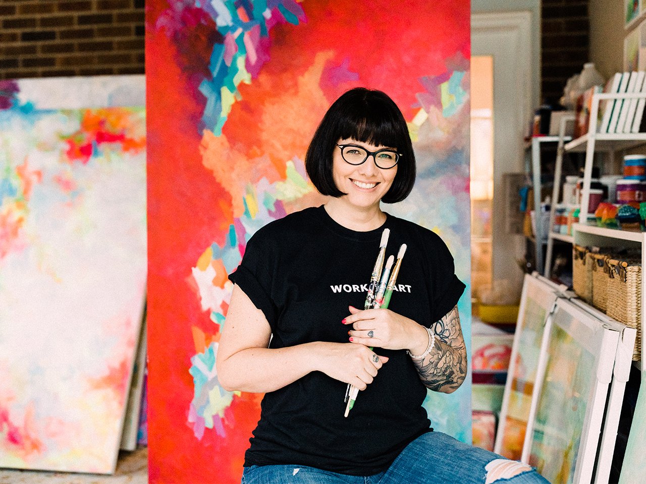 Canadian artist Sara Purves holding brushes in front of several large-scale paintings to illustrate an article on female artists in Canada.