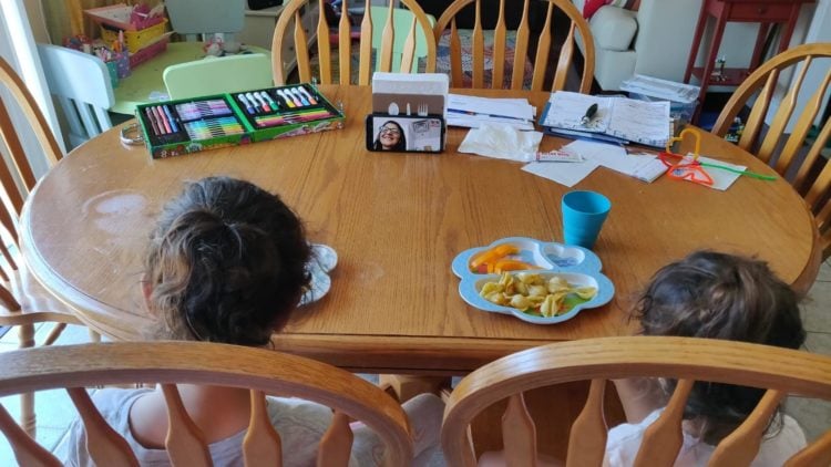 two small children sit at a kitchen table conferencing with an adult via zoom