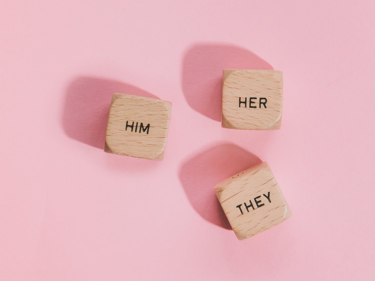 photo of three dice with "he," "she," and "they" written on top against pink backdrop