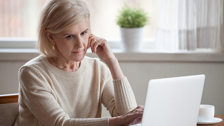 Frustrated grey hair sad middle aged woman sitting at table using computer.