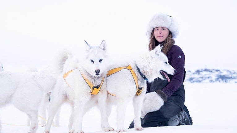 a woman in a parka and fur hat sits on a snowy field with three husky dogs