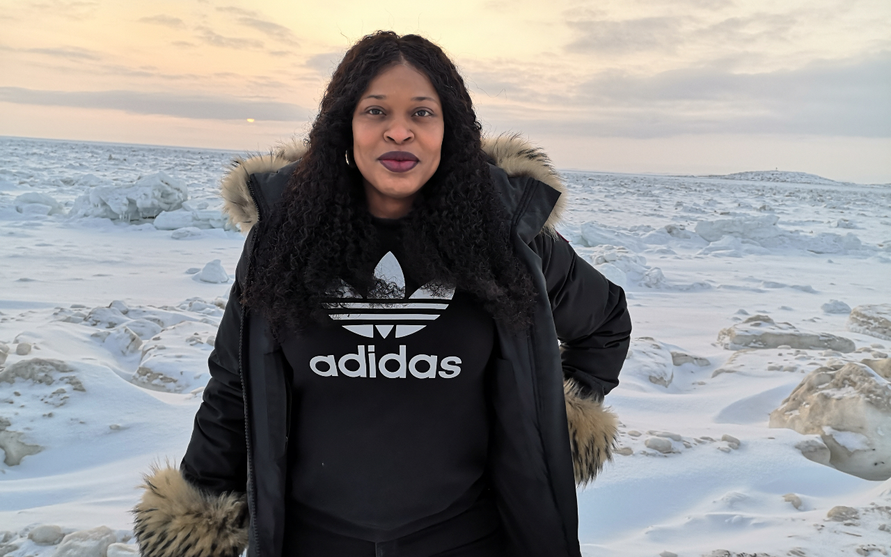 a woman in a black parka and adidas sweater stands on a snowy tundra near sunset