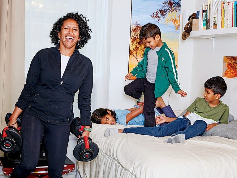 A mom holding weights standing and laughing; to her left, her three kids are on a bed.