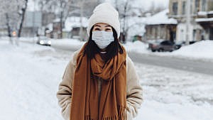 Woman stands on the snow, wearing a mask, hat and a scarf.