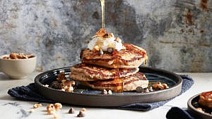 A stack of banana bread pancakes topped by whipped cream on a grey plate with maple syrup being drizzled on them