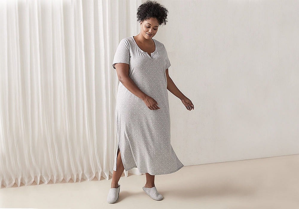 Penningtons Feathered Short Sleeve Sleepshirt. Woman swaying her arms to the side wearing a grey long dress with light white dots throughout.