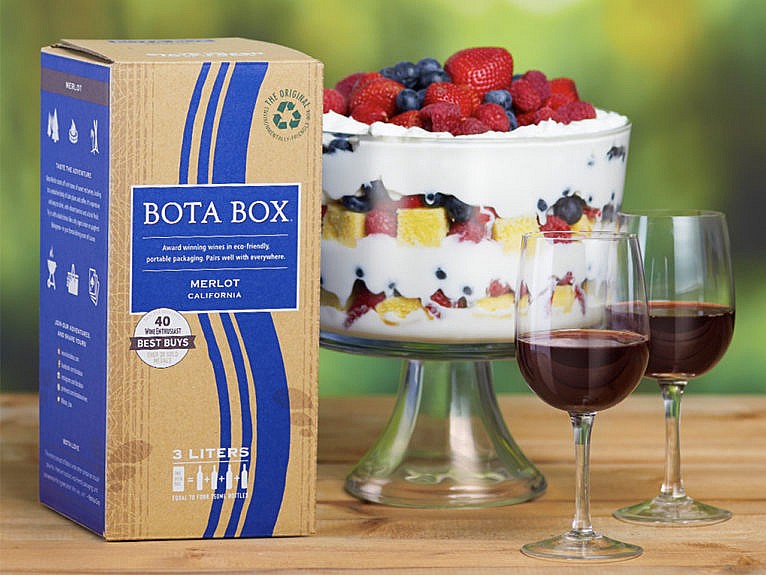 A box of wine sits on a table outdoors next to two glasses of red wine and a dessert triffle.