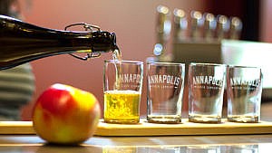 A bottle of Annapolis cider pouring into a flight of cider glasses, with an apple in the foreground