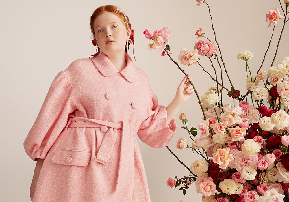 A woman in a pink Simone Rocha x H&M coat next to a large vase of roses.