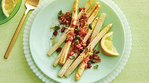 White asparagus with bacon, italian bread crumbs, parsley, lemon zest, browned butter and a lemon wedge on a green plate.