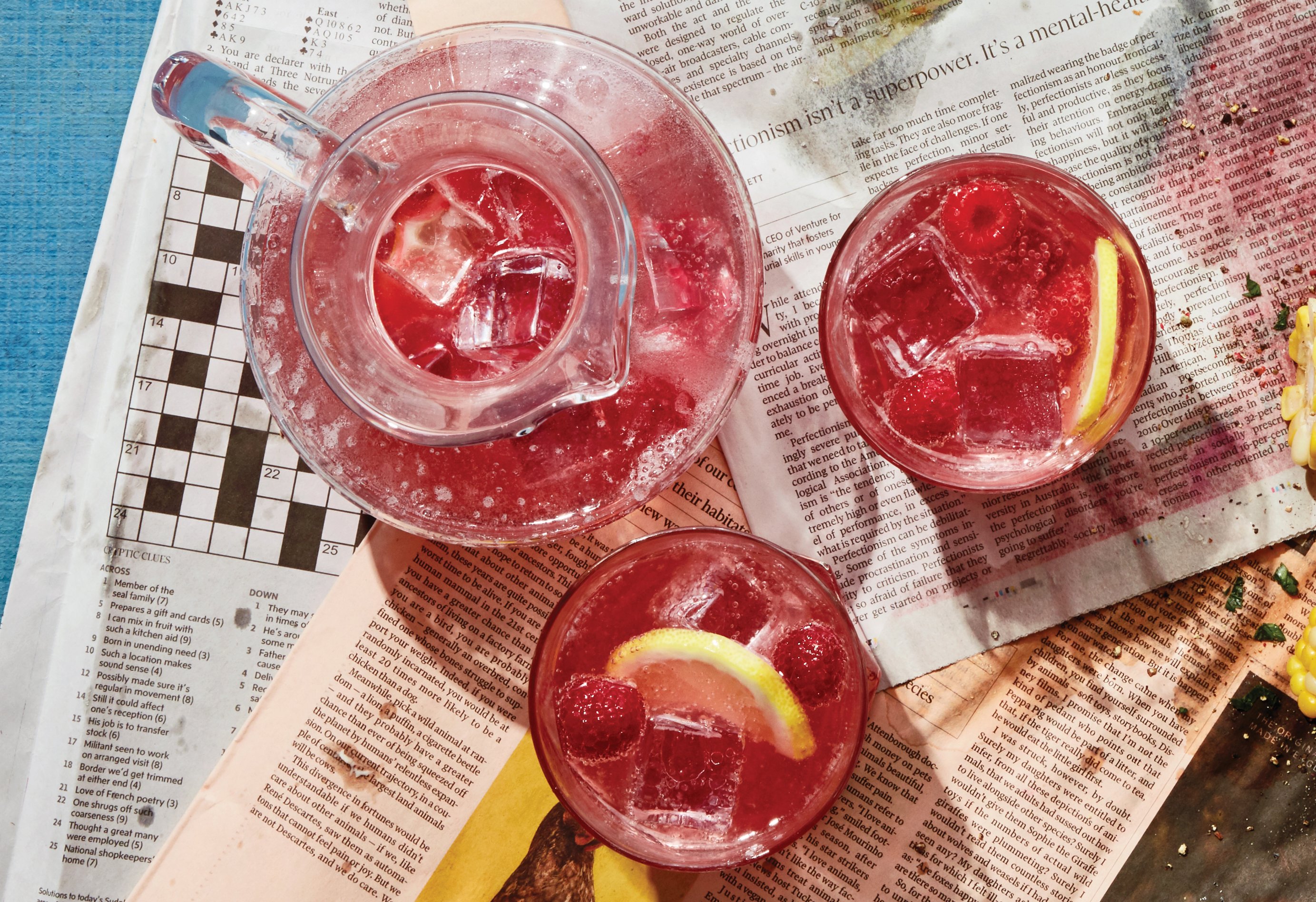 A pitcher of ice-filled raspberry cordial and two glasses, garnished with lemons, on a newspaper backdrop