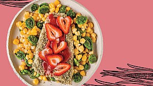 A piece of breaded pickerel topped with strawberries in a bed of hominy and fiddleheads on an off-white ceramic plate