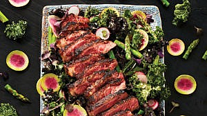 Broiled steak and spring kale caesar salad on a square platter surrounded by asparagus and radishes