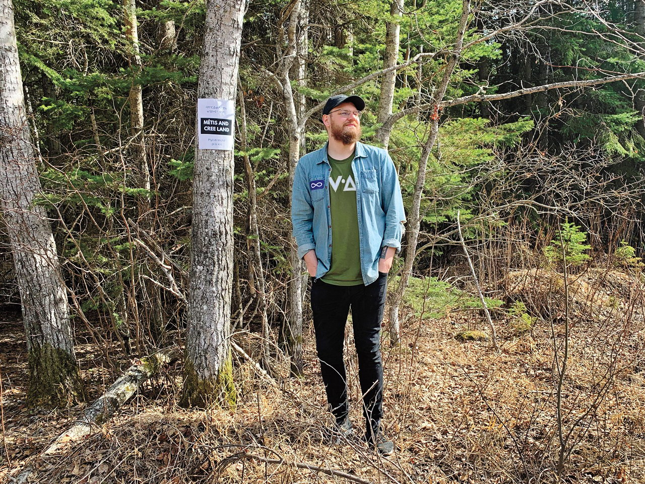 An Michif napew man in a denim shirt and black pants standing in the forest